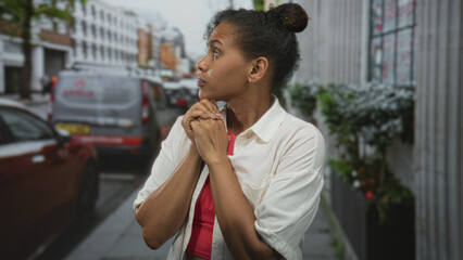 Woman clasping hands to chest on a busy city street, eyes closed and lips pursed, white shirt and...