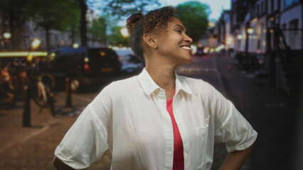 Woman smiling with hands on hips wearing white shirt and red top on a city street under lampposts...