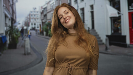 Fototapeta premium Smiling redheaded woman raises forearms on busy urban street lined with buildings; celebration joy.