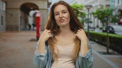 Caucasian young woman touches hair and flips strands in front of building in daylight; insecurity doubt.