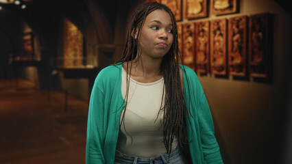 Woman clasps hands under chin inside building near framed art, braided hair, green cardigan and...