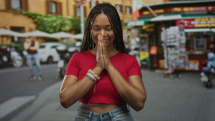 Woman with hands pressed together praying on a city street near market stalls and parked scooters, red crop top and bracelets visible; gratitude peace.
