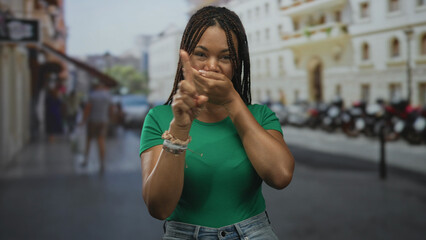 Fototapeta premium Young african american woman with braided hair covering mouth with both hands on urban street near building; surprise secrecy.