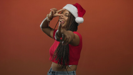 Young african american woman wearing santa hat and red crop top pointing both fingers at camera in studio with orange backdrop; playful.