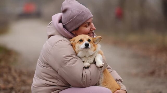 A woman in a hat and down jacket tenderly hugs a happy corgi on an autumn walk. Human-dog friendship.