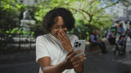 Young african american woman smiling while holding smartphone with hand on chest in lush green park; connection joy.
