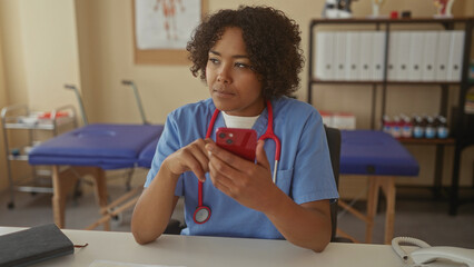 Woman in blue scrubs wearing stethoscope sits at desk taps red smartphone in clinic room; professional focus calm determination.