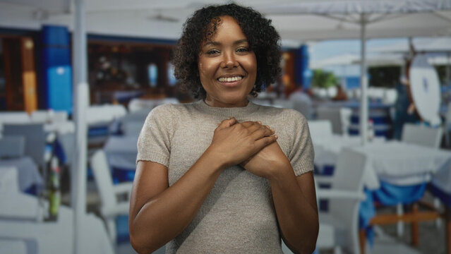 Young african american woman with curly hair places hands on chest in outdoor restaurant building while smiling; gratitude.