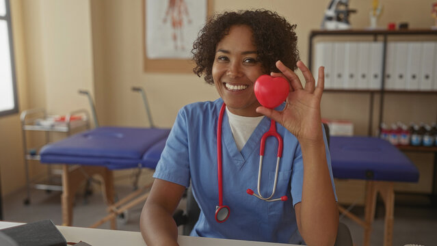 Young african american woman doctor smiles while holding red heart model in clinic; heart health compassion care.