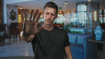 Man in black t shirt with hand palm forward to camera stop in hotel building lobby; defiance boundary privacy.