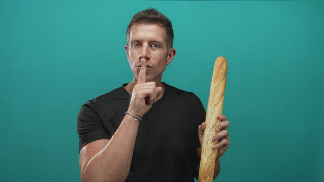 Man holding baguette and finger to lips for silence in teal studio with casual black t shirt and bracelet visible; silence.