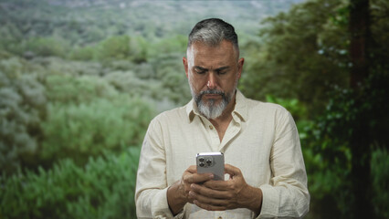 Man in linen shirt looking down at smartphone and holding it with both hands in a forest setting among green bushes and grassy field; concentration.