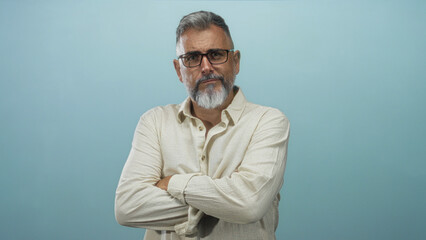 Man middle age with folded arms and glasses in a light blue studio, looking at camera; confidence...