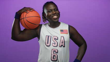 Man holding basketball on shoulder wearing usa jersey and wristband in purple studio smiling and posing; joy athleticism.