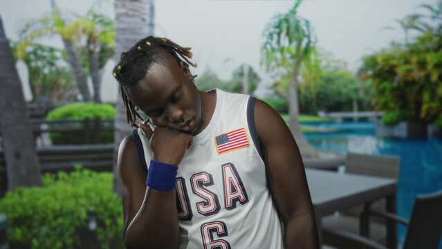 Man with hand on chin wearing usa jersey by resort pool patio table and palm trees; quiet contemplation. - Powered by Adobe