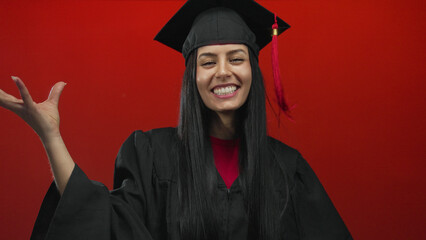 Hispanic woman celebrating graduation in a cap and gown over a vibrant red background with a joyful expression and raised hand.