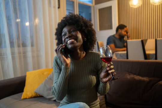 Young African American woman talking on phone while drinking wine at home in evening, smiling