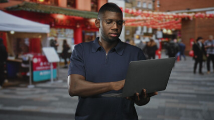 Fototapeta premium Young black man wearing glasses typing on laptop, hands visible in busy street market; concentration productivity focus.
