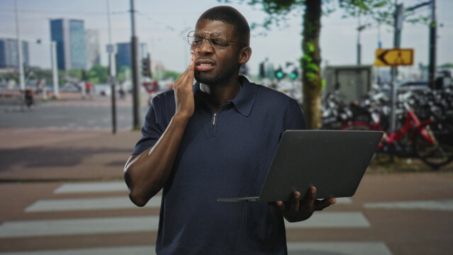 Young man holding laptop, hand on cheek wincing at a busy street crosswalk with bicycles and traffic lights; remote work difficulty pain.