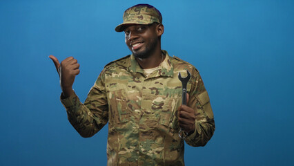 Fototapeta premium Man in camouflage uniform and cap holding wrench and pointing thumb to left in studio; confidence teamwork.