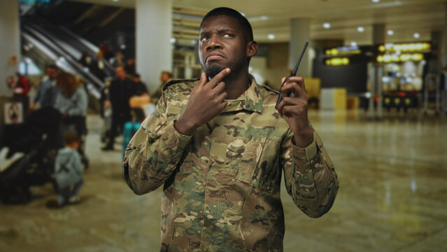 Soldier touching chin while holding radio in airport terminal, camouflage uniform visible and face focused upward; concern duty.