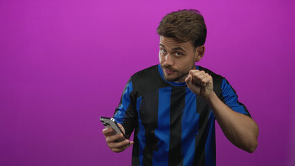 Young hispanic man holding smartphone and making a small fist gesture while looking skeptically at the screen in a studio with magenta backdrop; skepticism.