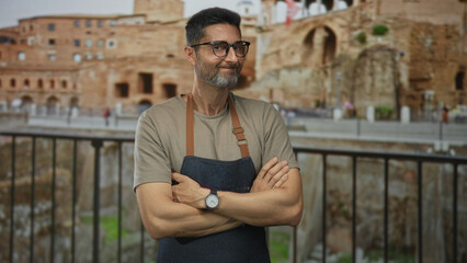 Man with arms crossed wearing denim apron at building ruins by ancient stone walls smiling to camera; confidence craft.