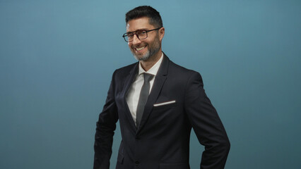 Man in dark suit smiling with glasses and hands on hips in studio against teal backdrop; confidence...