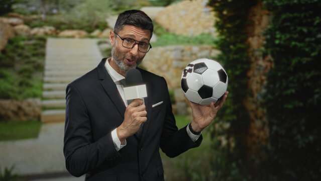 Man reporter holding microphone and soccer ball at building steps wearing suit and glasses; sports broadcast excitement.