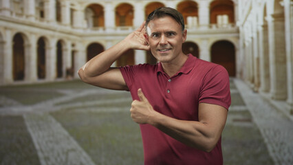 Man holds hand to ear and gives thumbs up in a historic courtyard building with arched colonnade,...