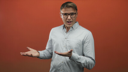 Man with open palms gesturing in studio, wearing light buttondown shirt and glasses, standing against orange backdrop, facing left; confusion questioning.