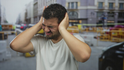 Fototapeta premium Young hispanic man wearing white tshirt pressing hands to ear on busy construction site street; stress.