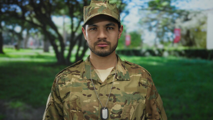 Man wearing green camouflage uniform, cap, and dogtags looks at camera while standing in a forest; duty.