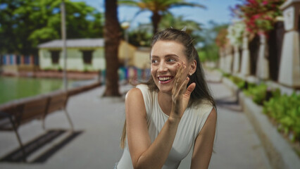 Woman with hand to cheek smiling and laughing on a sunny street by a green park walkway with bench and palm tree; happiness playfulness.
