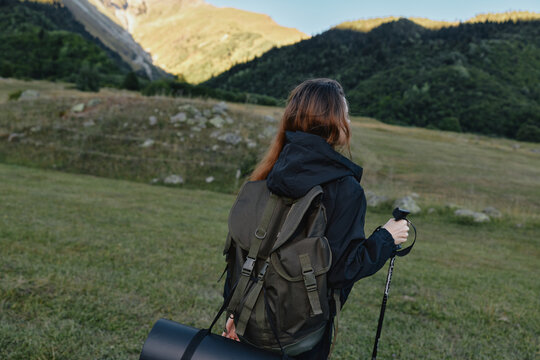 A lone hiker stands in a wide alpine meadow, wearing a backpack and using a trekking pole. She gazes toward distant mountains, exuding calm focus and adventurous spirit.