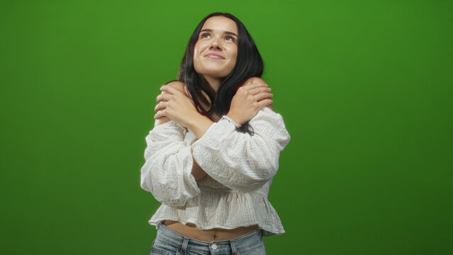 Woman hugging shoulders with hands in studio against green screen, wearing white blouse and jeans; self care contentment. - Powered by Adobe