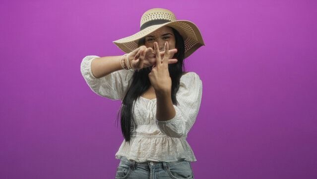 Woman with straw hat showing hands, forms hashtag with fingers in studio against purple backdrop; joyful playfulness summer.