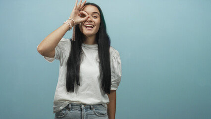 Woman making ok sign around eye with hand, smiling, long black hair, bracelets, white t shirt and jeans in studio against blue backdrop; joyful playfulness.