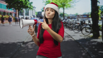 Woman in santa hat points finger to megaphone on street, wearing red shirt and smiling broadly; festive announcement.