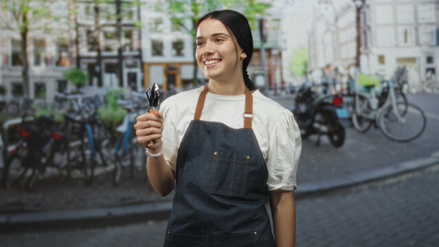 Woman gardener smiles holding small trowels in hand and wearing denim apron on cobblestone street with parked bicycles; contentment.
