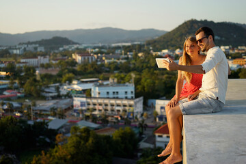 A happy couple poses for a tablet selfie on a rooftop with a city and mountain view during a beautiful sunset, capturing the warmth of the moment and scenic surroundings.