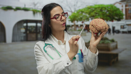 Young hispanic woman doctor with stethoscope holds model brain on street demonstrating medical concept outdoors.
