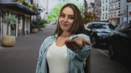Fototapeta premium Woman with long hair smiling, arm extended and hand reaching toward camera on a city street; confidence invitation.