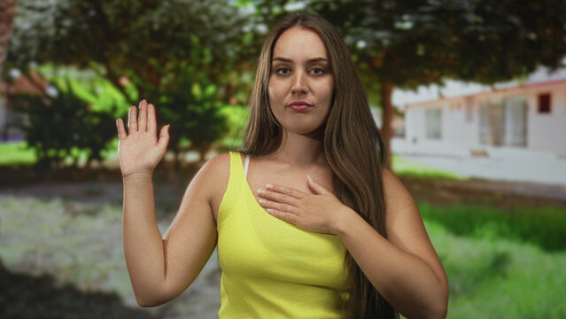 Woman with one hand raised and other hand pressed to bare chest beside a residential building and shaded trees; sincerity.