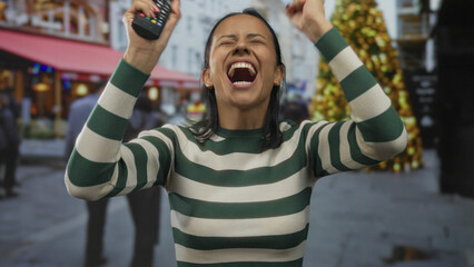 Woman smiling with eyes closed holding remote outdoors near festive christmas market in winter urban street scene captured mid-motion showing joy and happiness