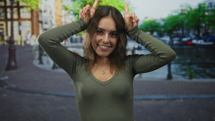 Woman with hands raised making horns gesture on amsterdam street near canal, smiling and showing fingers; playful joy.