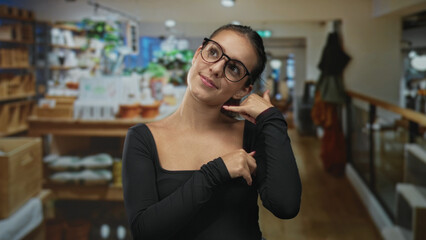 Woman with exposed shoulder wearing glasses making a call gesture in a home decor shop interior among wooden shelves and displays; playful curiosity.
