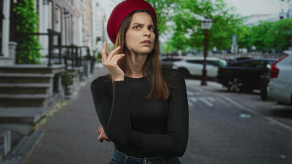 Fototapeta premium Woman touching red beret on street with hand to head, frowning and crossing one arm over waist while wearing black shirt and jeans; urban confusion.