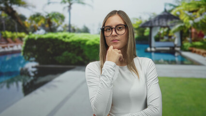 Young hispanic woman with hand on chin and glasses at a poolside resort gazebo by a swimming pool wearing a white long sleeve shirt and crossed arms; thoughtful.