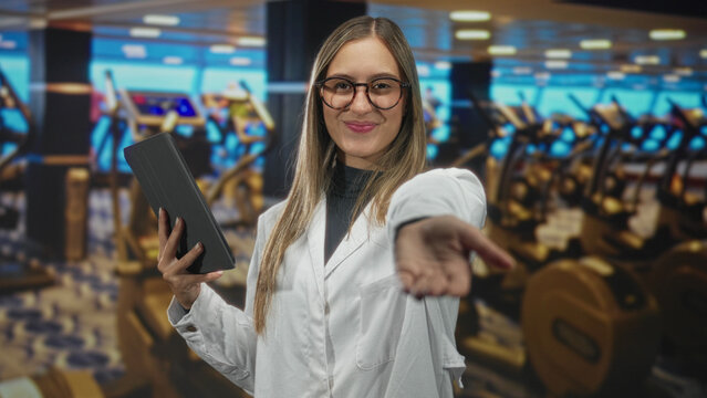 Woman in white coat holding a tablet, smiling with eyes closed and extending her hand toward camera in a gym with treadmills and dumbbells; confidence.